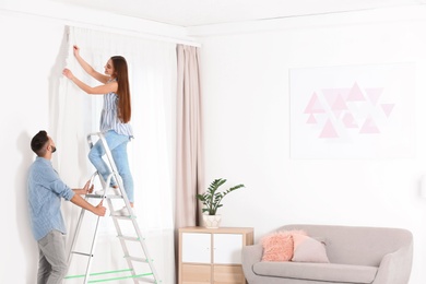 Young couple hanging window curtain in room. Space for text