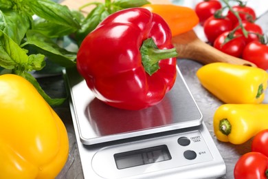 Photo of Kitchen scale with bell pepper among basil and tomatoes on grey textured table, closeup