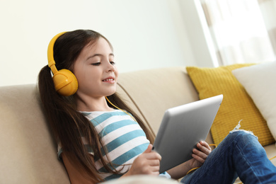 Photo of Cute little girl with headphones and tablet listening to audiobook at home