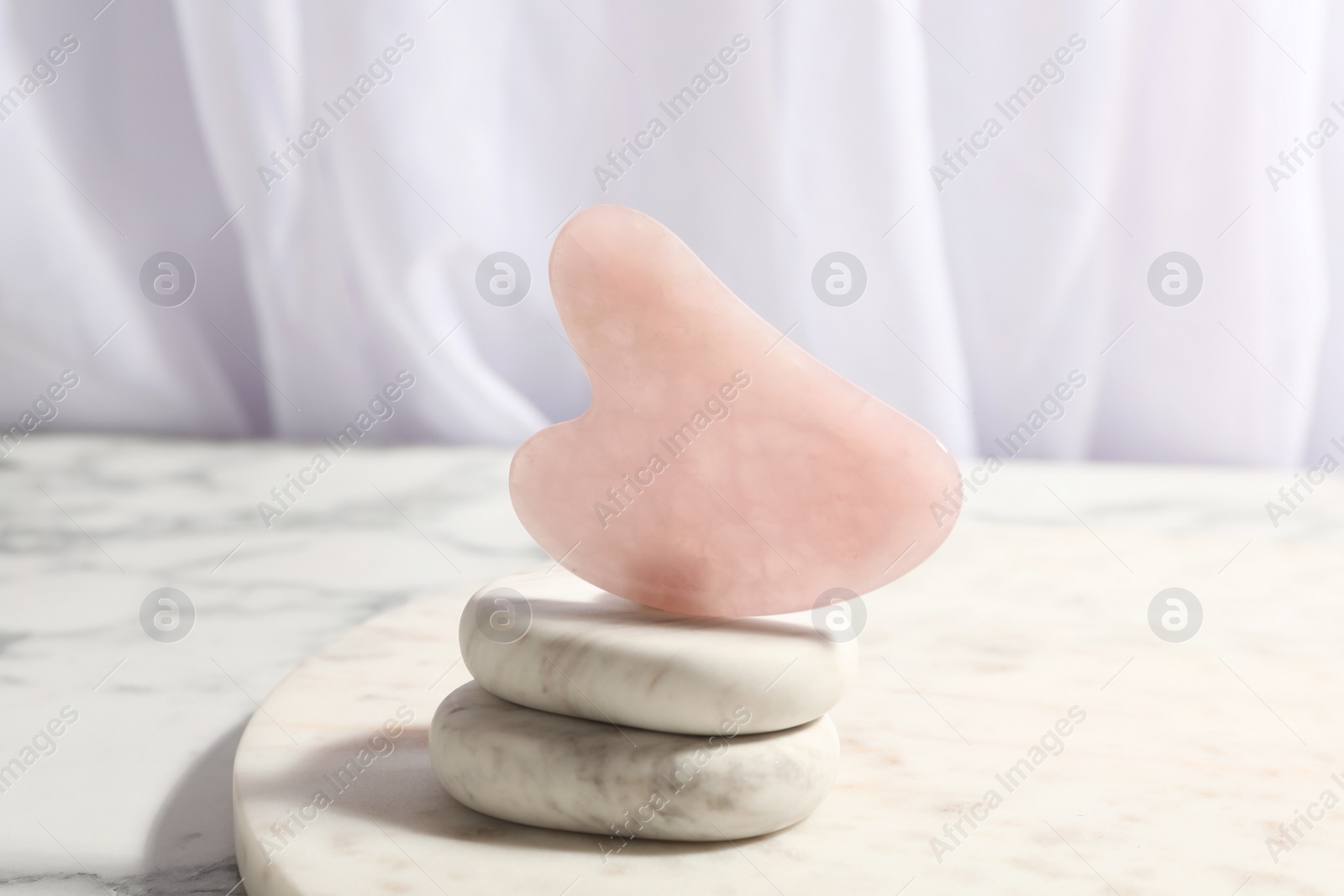 Photo of Rose quartz gua sha tool and spa stones on white marble table