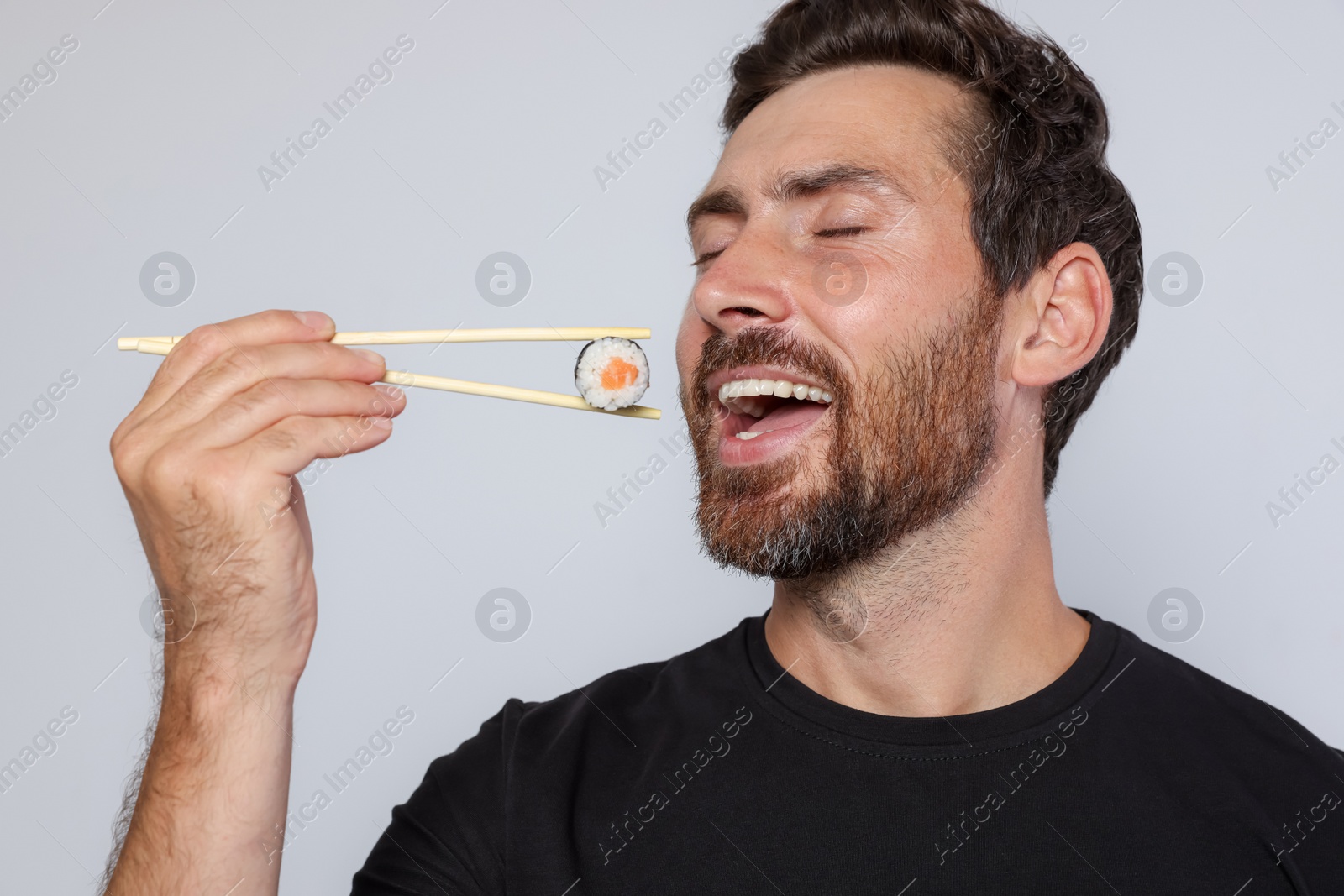 Photo of Happy man eating sushi roll with chopsticks on light grey background, closeup