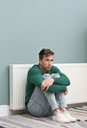 Sad young man suffering from cold on floor near radiator