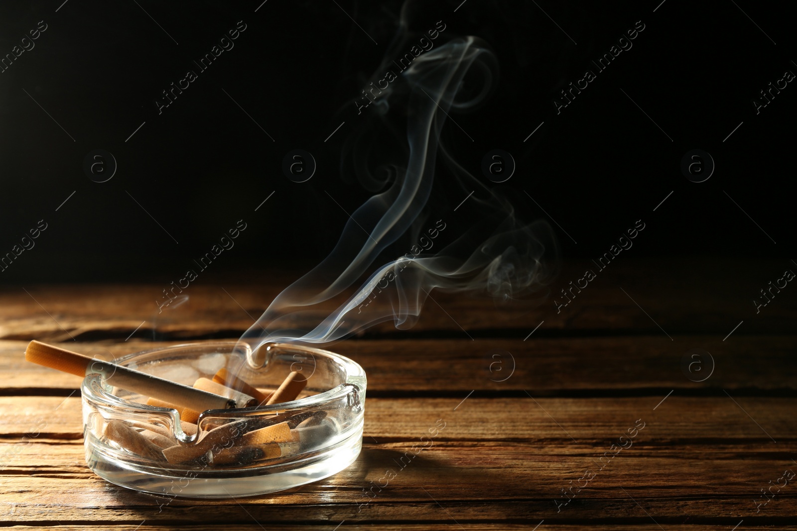 Photo of Smoldering cigarette in glass ashtray on wooden table against black background. Space for text