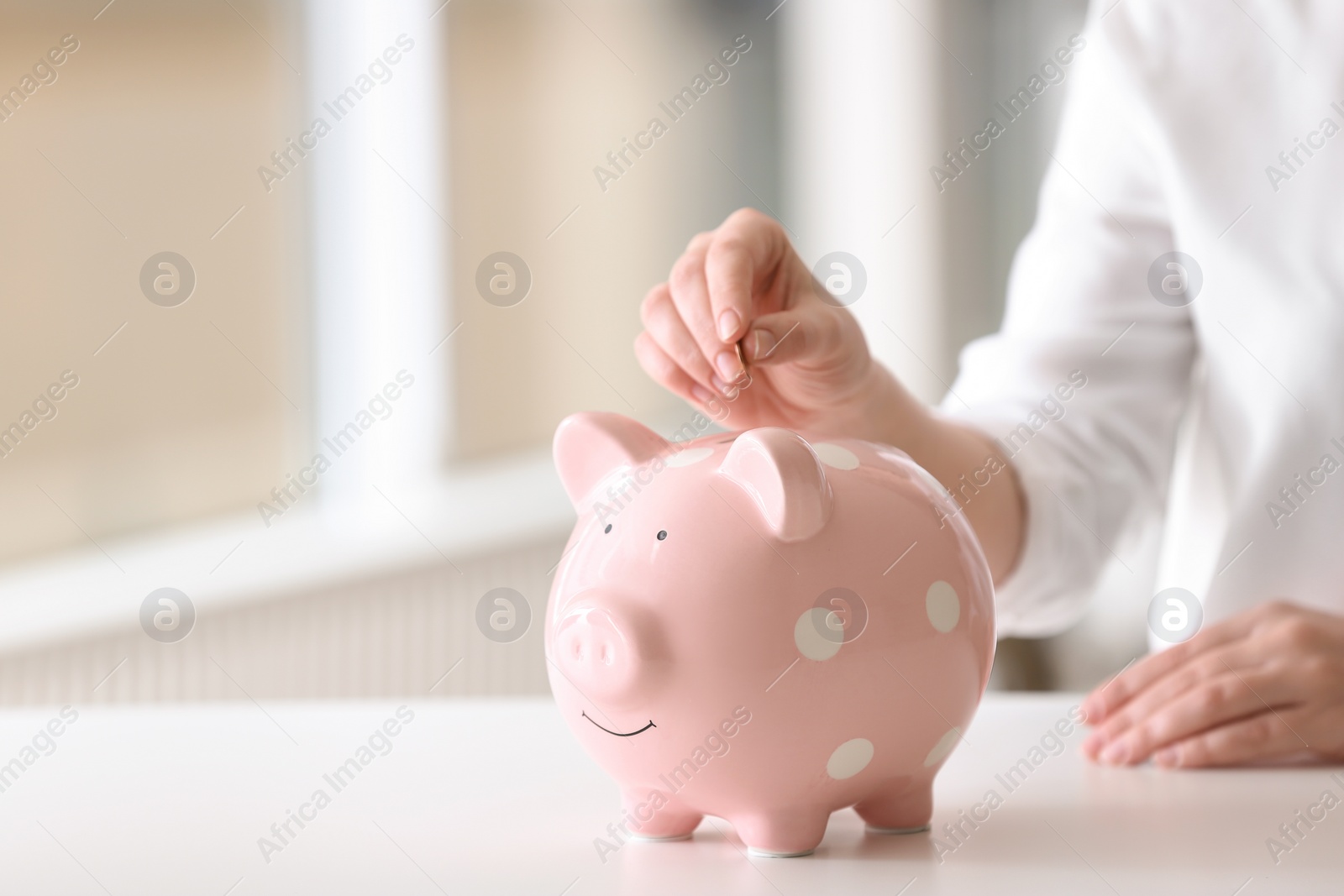 Photo of Woman putting coin into piggy bank at table indoors, closeup. Space for text