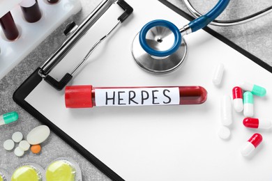 Test tube with word Herpes, different pills, clipboard and stethoscope on light grey table, flat lay