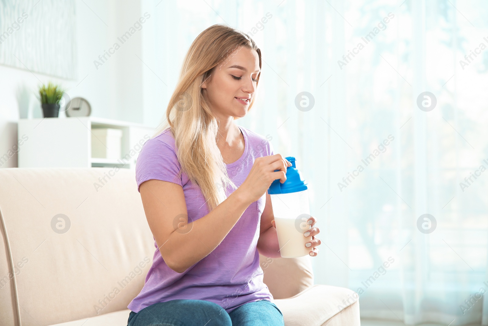 Photo of Young woman with bottle of protein shake sitting on sofa at home