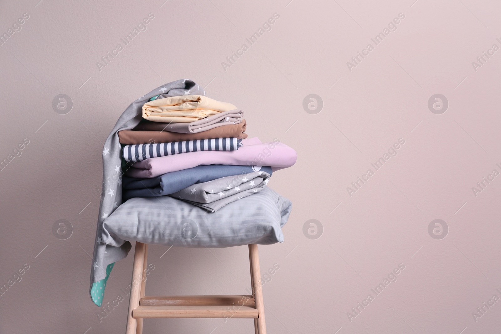 Photo of Stack of clean bed sheets and pillow on stool near beige wall. Space for text