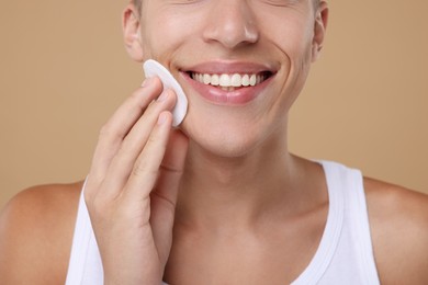 Man cleaning face with cotton pad on beige background, closeup