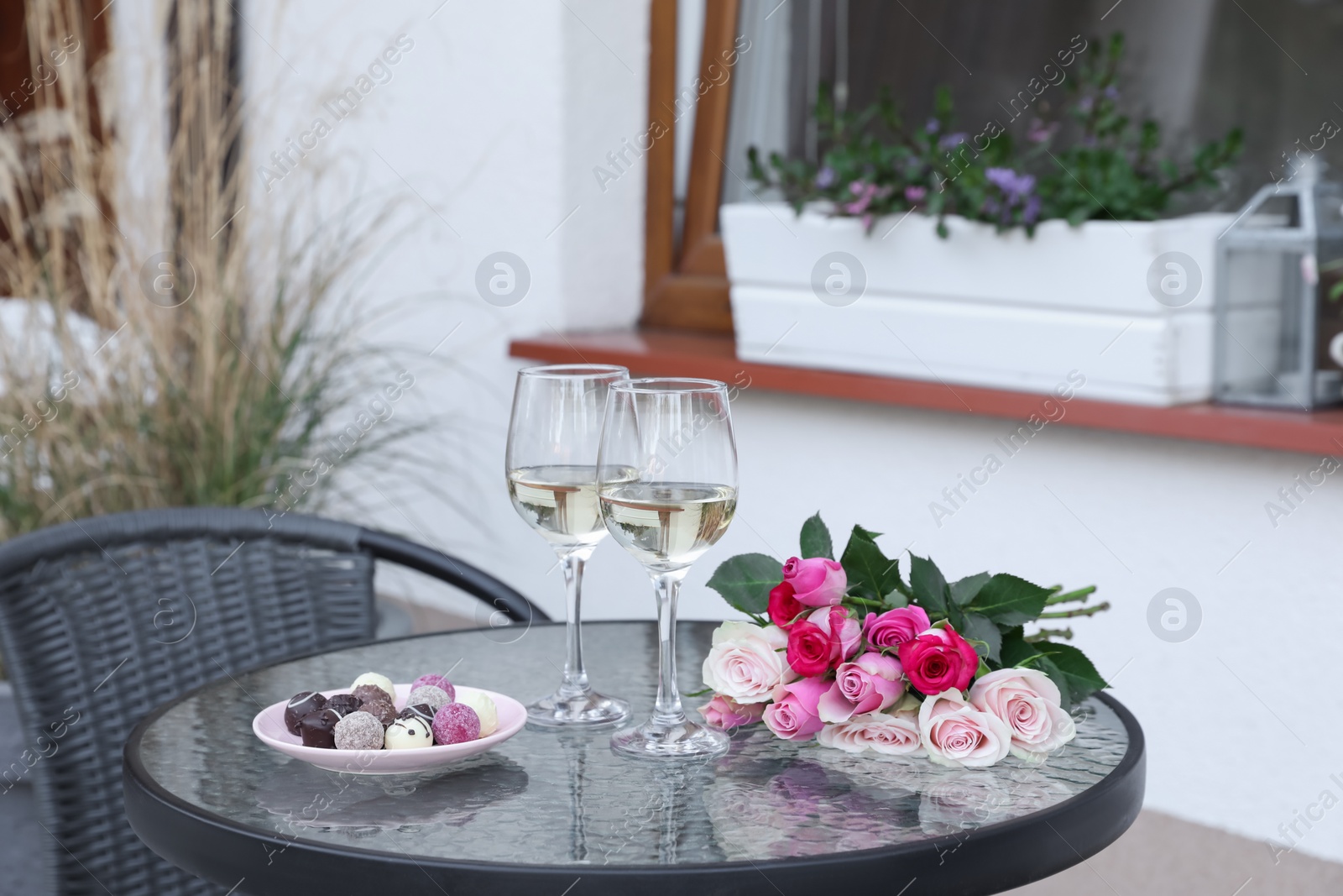 Photo of Bouquet of roses, glasses with wine and candies on glass table on outdoor terrace