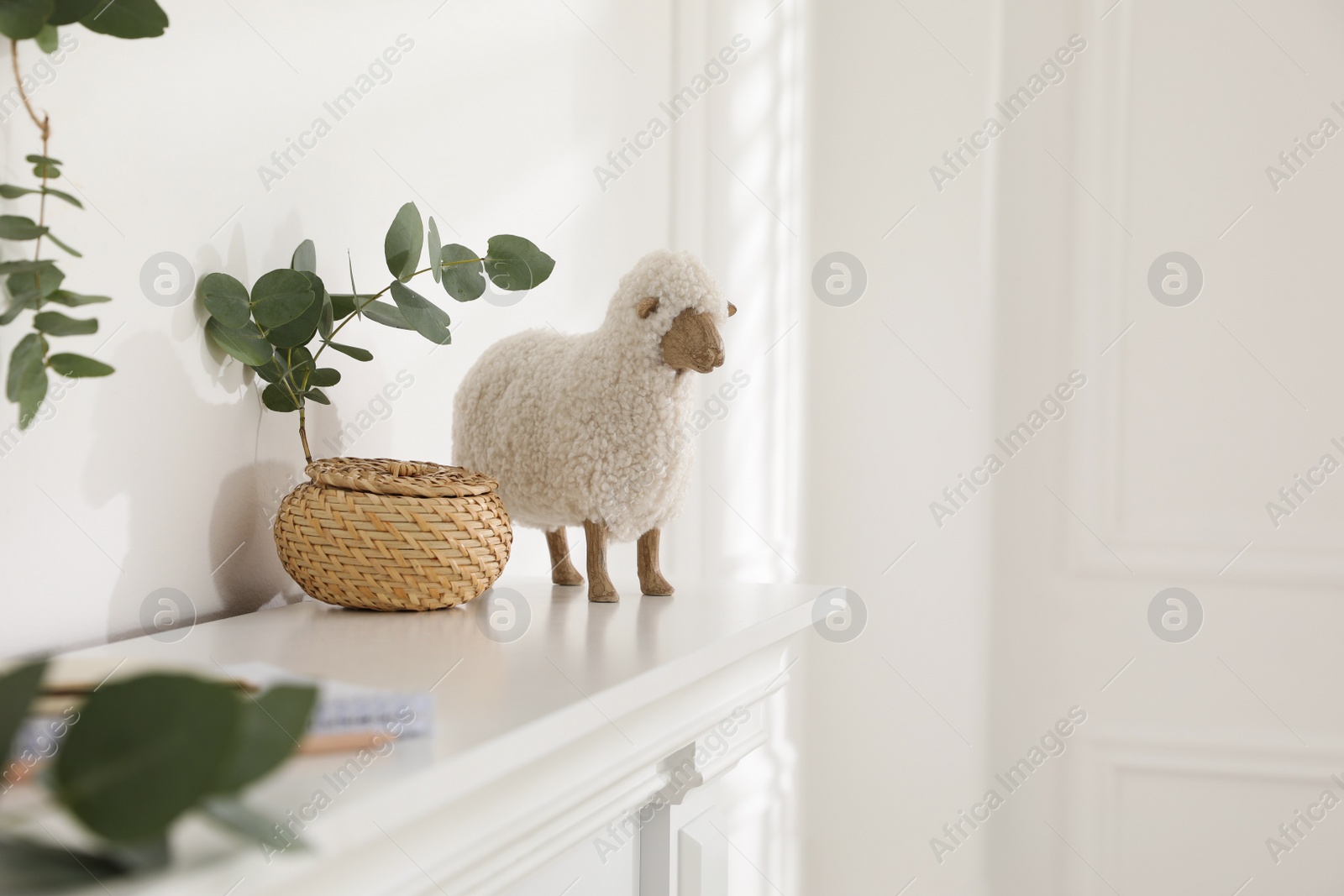 Photo of Wicker box with eucalyptus branch and decorative sheep on white shelf indoors, space for text