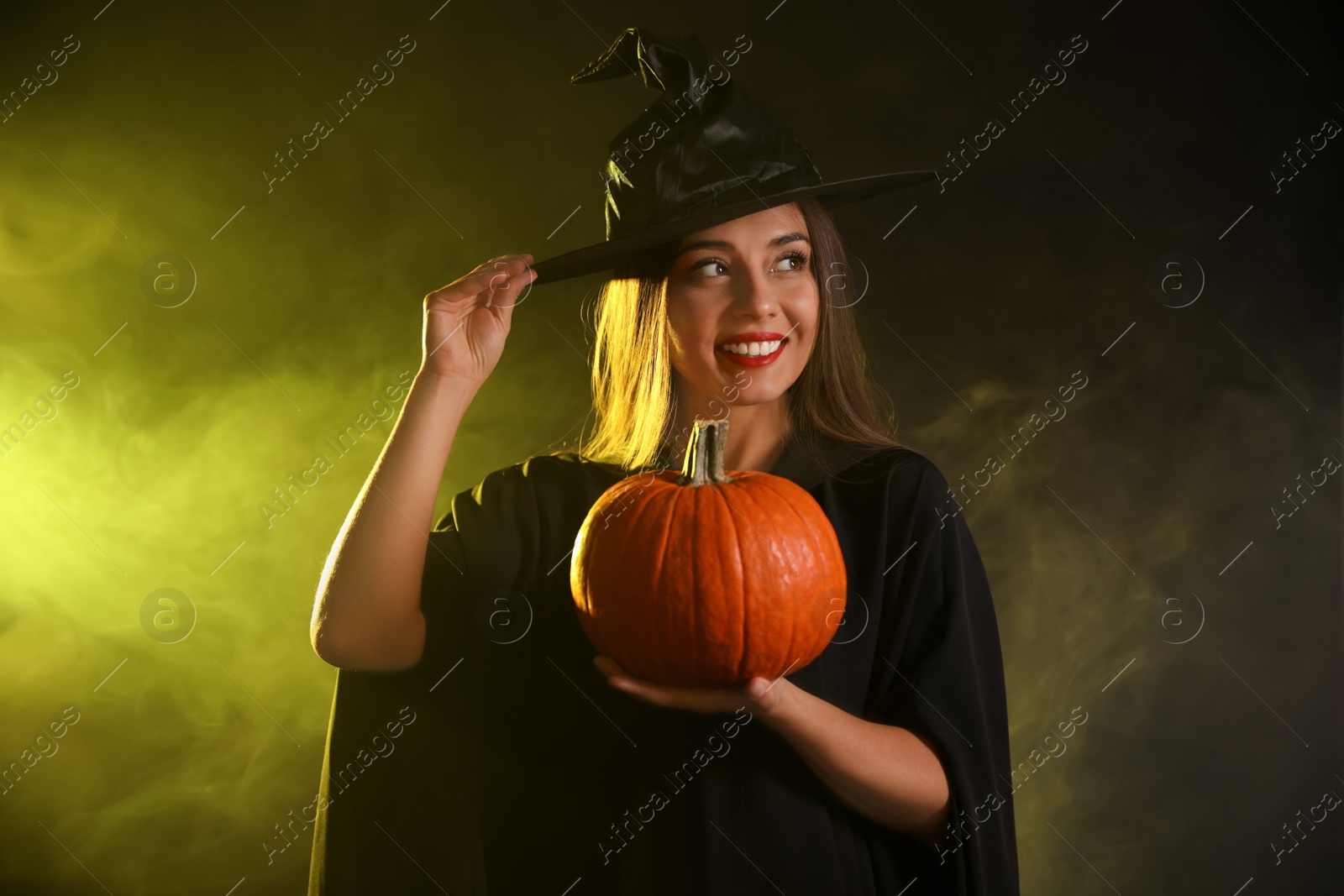 Photo of Young woman wearing witch costume with pumpkin in smoke cloud on dark background. Halloween party