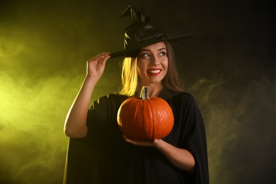 Young woman wearing witch costume with pumpkin in smoke cloud on dark background. Halloween party