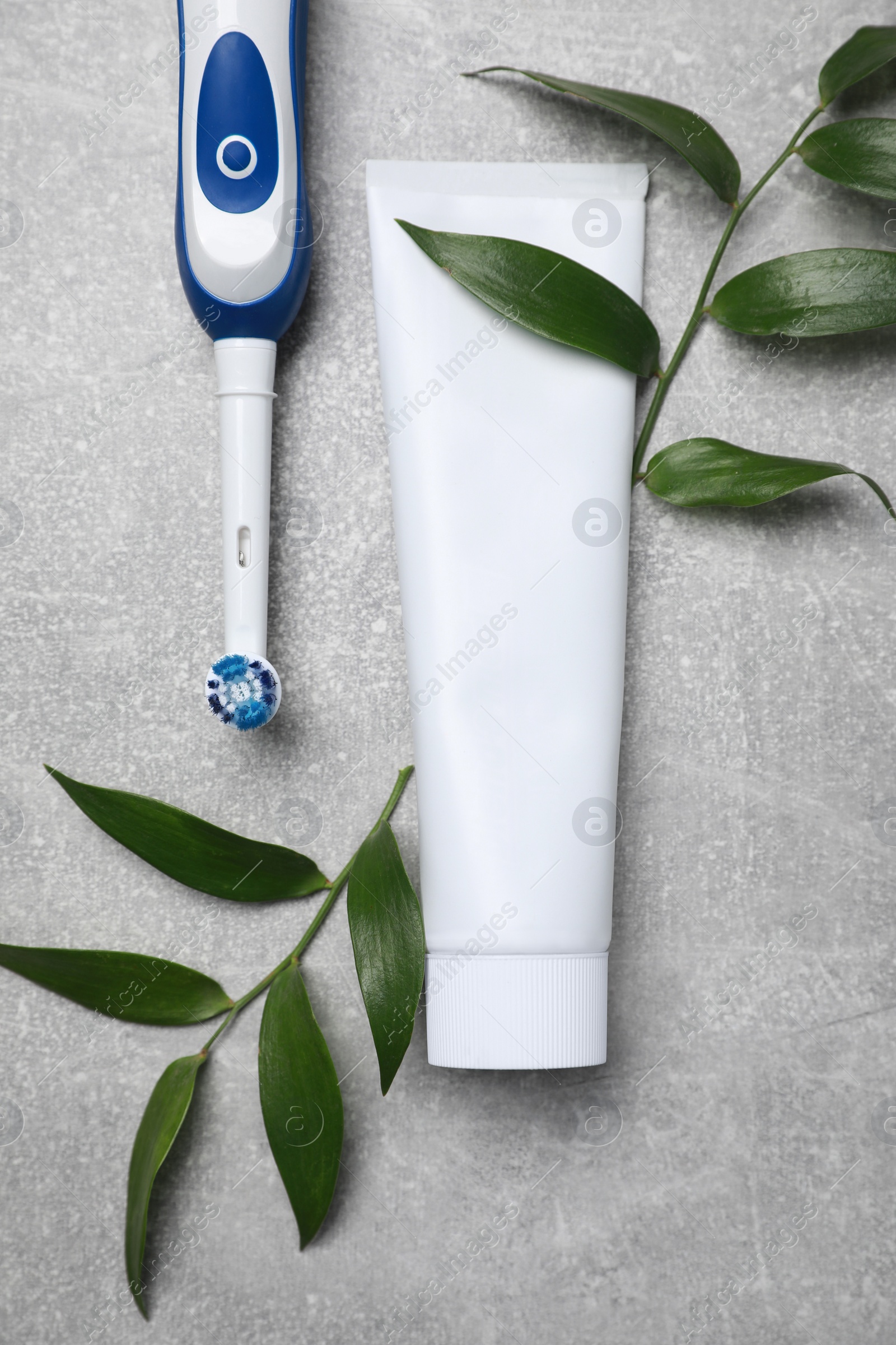 Photo of Electric toothbrush, tube of paste and green leaves on grey textured table, flat lay