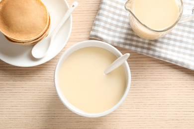 Photo of Bowl with condensed milk and pancakes served on wooden table, top view. Dairy products