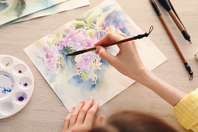 Photo of Woman painting flowers with watercolor at white wooden table, above view. Creative artwork