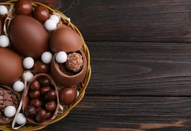 Photo of Tasty chocolate eggs and sweets in wicker basket on wooden table, top view. Space for text