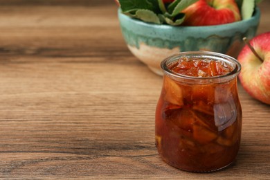 Photo of Tasty apple jam in glass jar and fresh fruits on wooden table, space for text