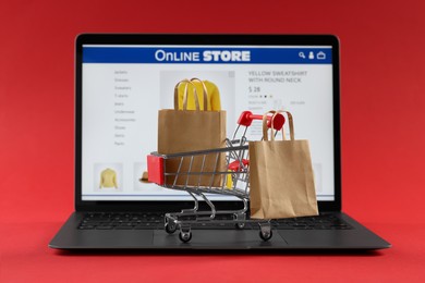 Photo of Online store. Laptop, mini shopping cart and paper bags on red background