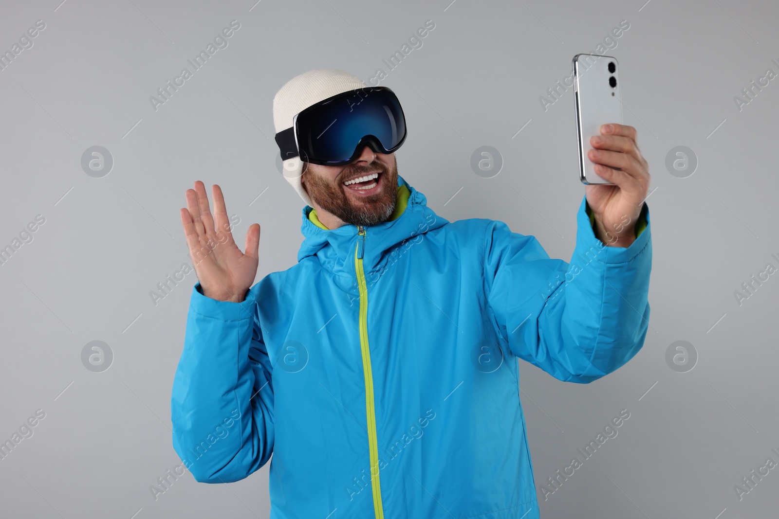 Photo of Winter sports. Happy man in ski suit and goggles taking selfie on gray background