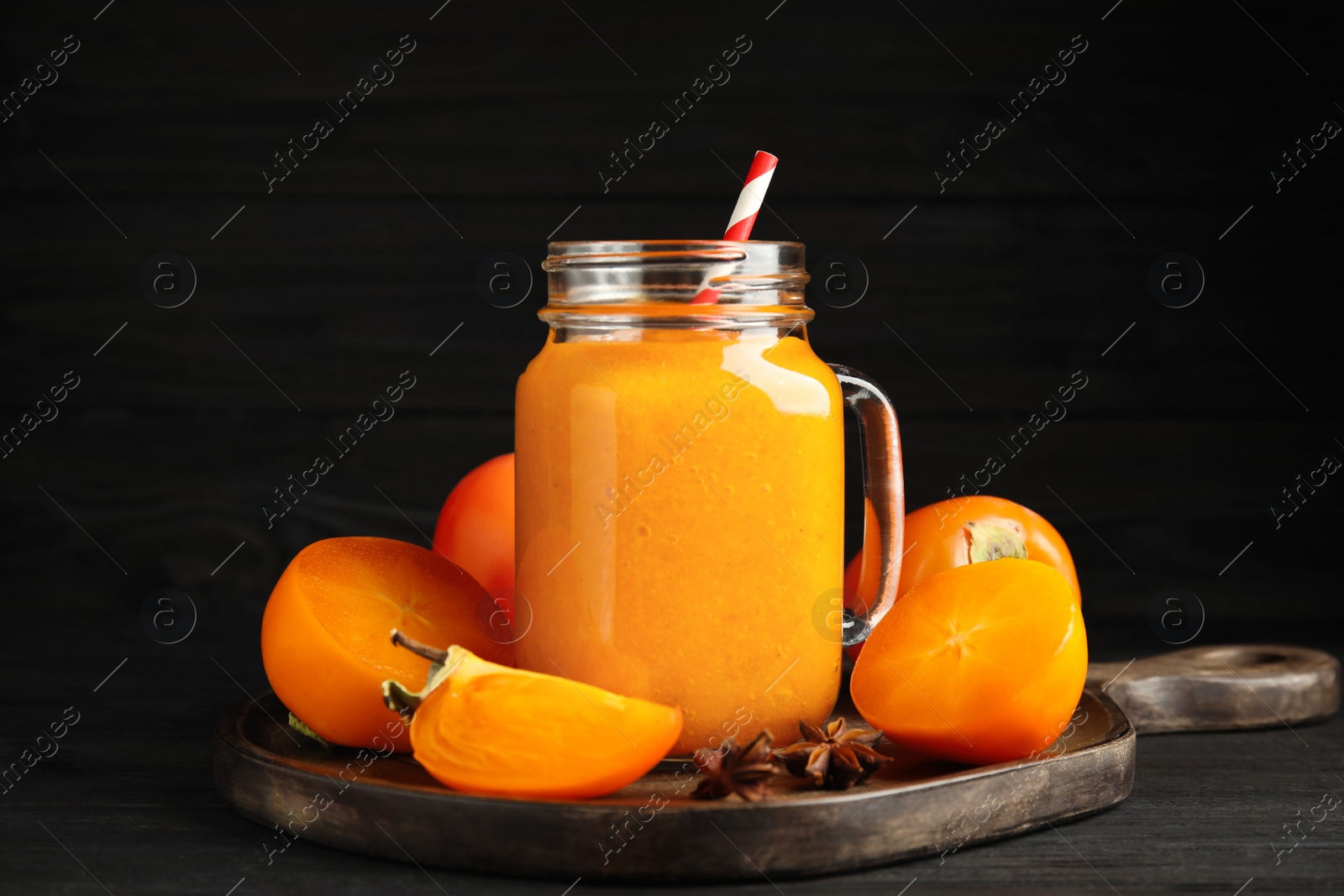 Photo of Tasty persimmon smoothie with straw and fresh fruits on black wooden table against dark background