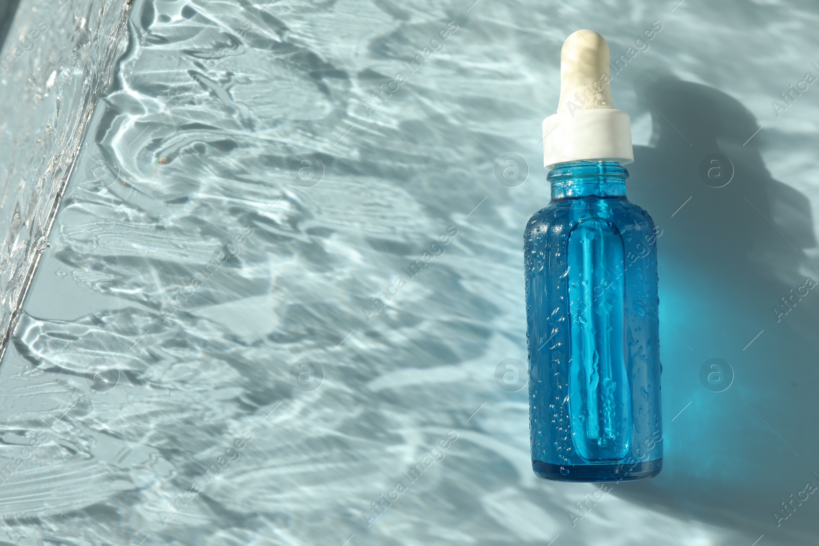 Photo of Wet bottle of cosmetic serum on light blue background, top view