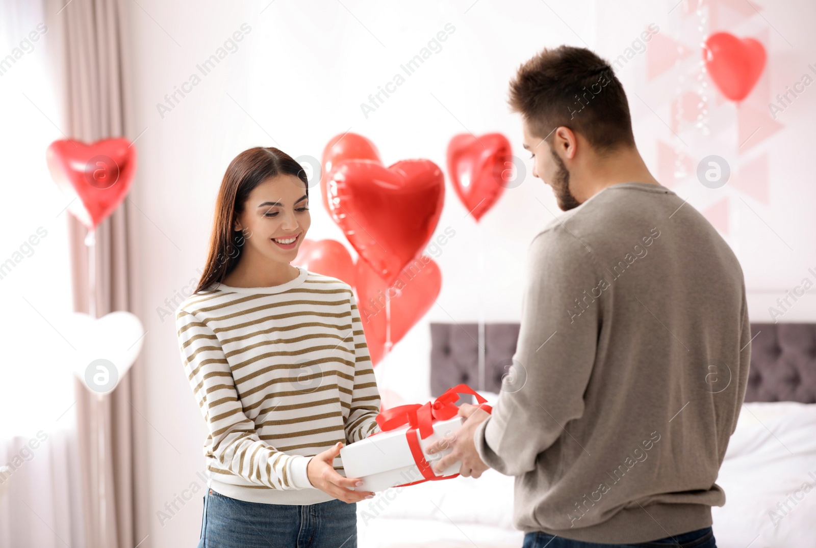 Photo of Young man presenting gift to his girlfriend in bedroom decorated with heart shaped balloons. Valentine's day celebration