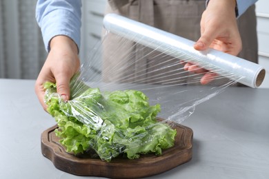 Woman putting plastic food wrap over fresh lettuce at light grey table indoors, closeup