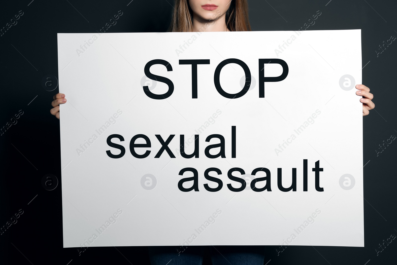Photo of Young woman holding card with words STOP SEXUAL ASSAULT against dark background, closeup