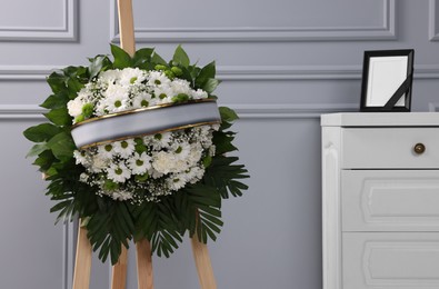 Photo of Funeral wreath of flowers on wooden stand and photo frame with black ribbon on white commode indoors