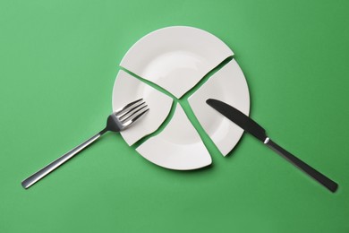 Photo of Pieces of broken ceramic plate and cutlery on green background, flat lay