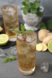 Photo of Glass of tasty ginger ale with ice cubes and ingredients on grey wooden table