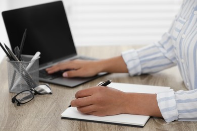 Photo of Woman writing notes while using laptop at wooden desk indoors, closeup