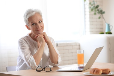 Photo of Mature woman with laptop sitting at table in kitchen. Smart aging