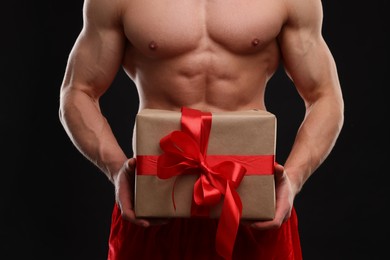 Attractive young man with muscular body holding Christmas gift box on black background, closeup