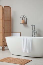 Photo of Modern ceramic bathtub and folding screen near white wall in room