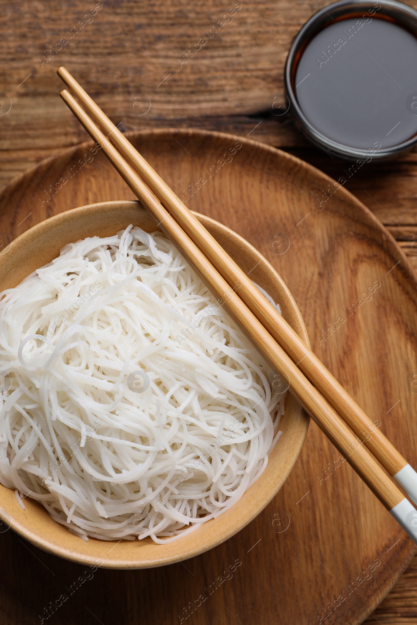 Photo of Bowl with cooked rice noodles, soy sauce and chopsticks on wooden table, flat lay