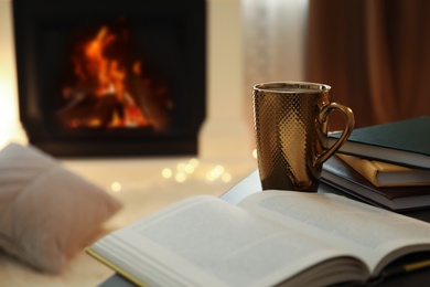 Photo of Cup of hot drink and book on table near fireplace at home. Cozy atmosphere