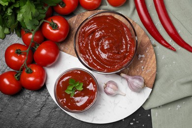 Photo of Organic ketchup in bowls and ingredients on black table, flat lay. Tomato sauce