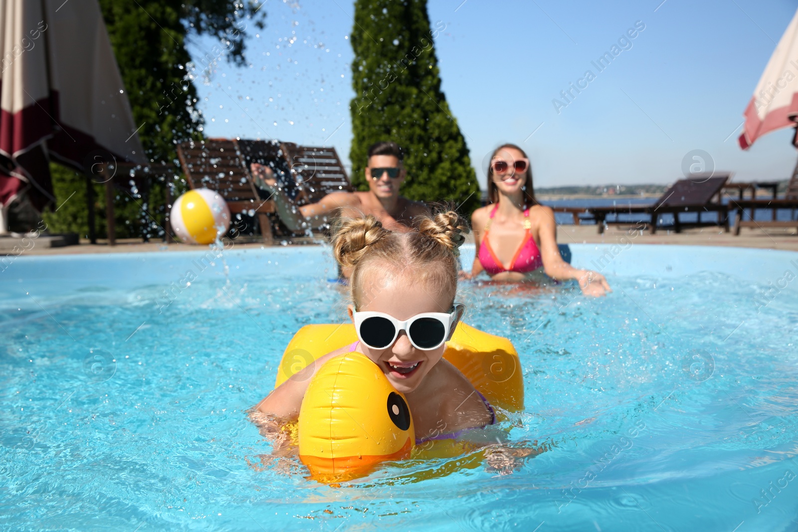 Photo of Little girl swimming with inflatable ring near her parents in outdoor pool on sunny summer day