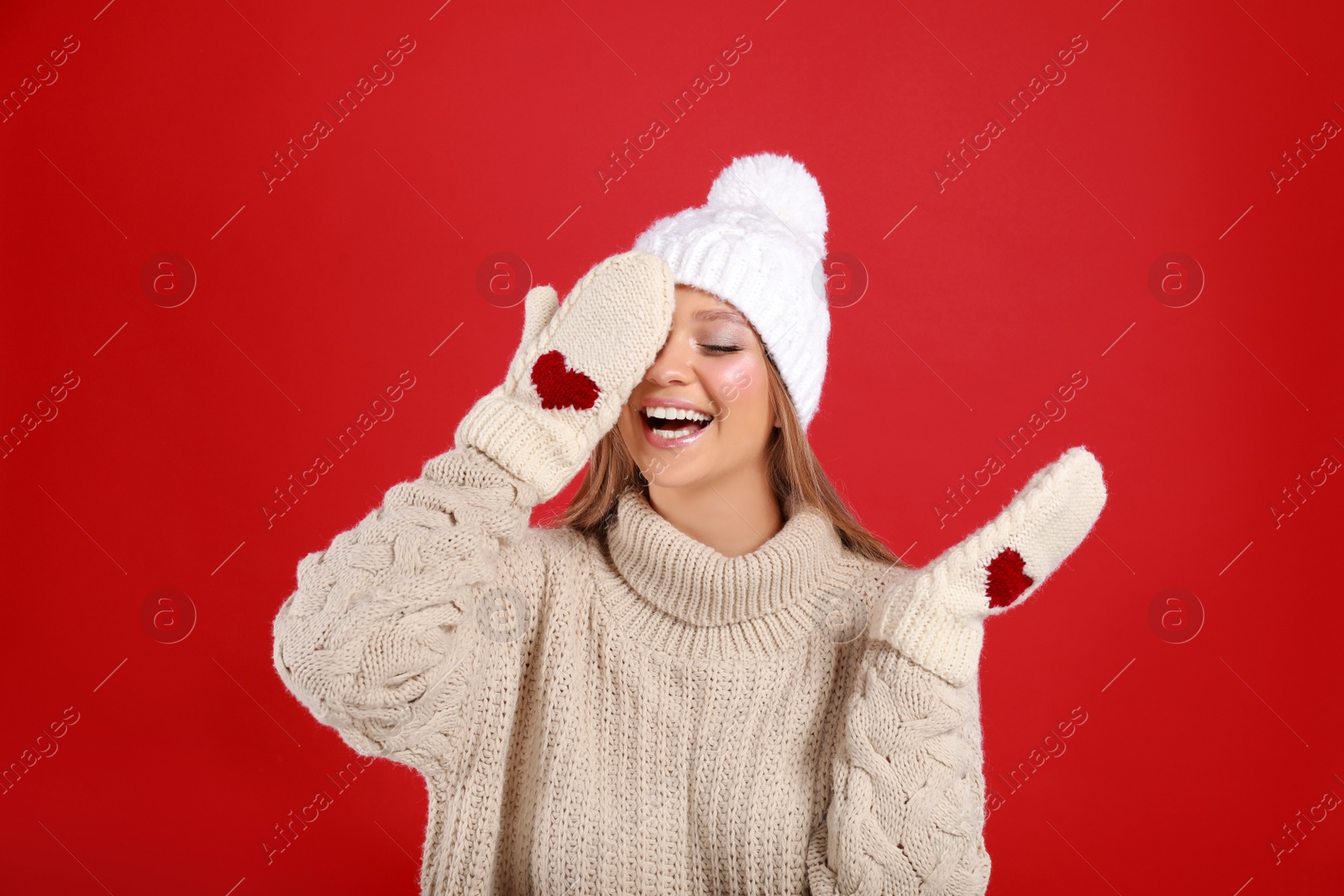 Photo of Funny young woman in warm sweater, mittens and hat on red background. Winter season