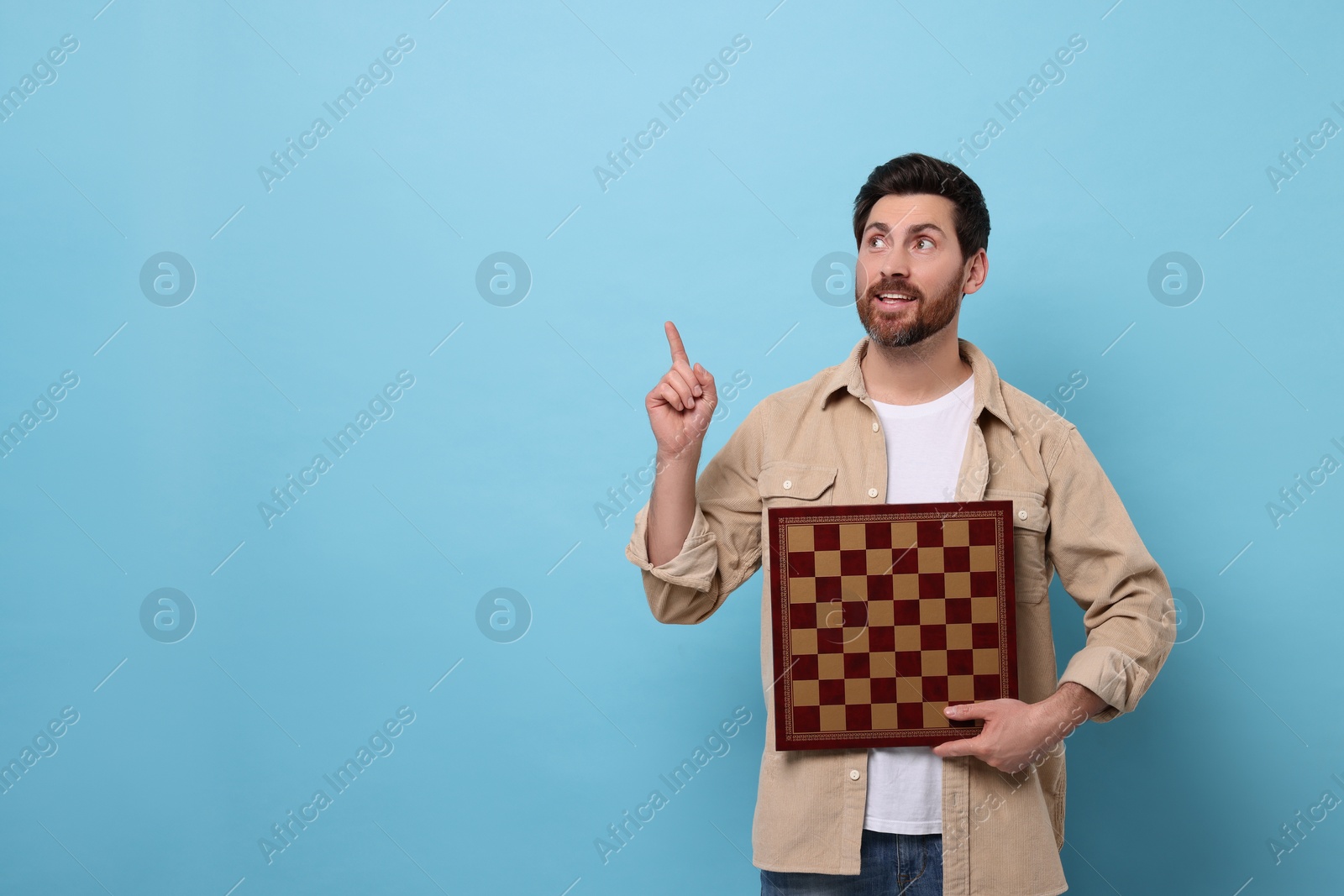 Photo of Smiling man holding chessboard and pointing at something on light blue background. Space for text