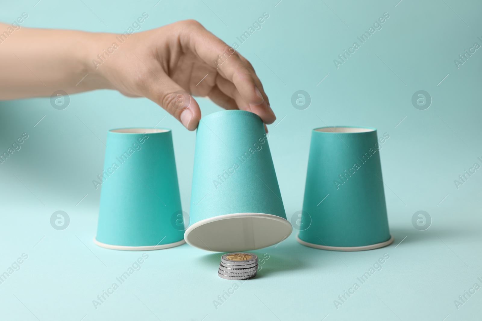 Photo of Woman showing coins under cup on light blue background, closeup. Thimblerig game