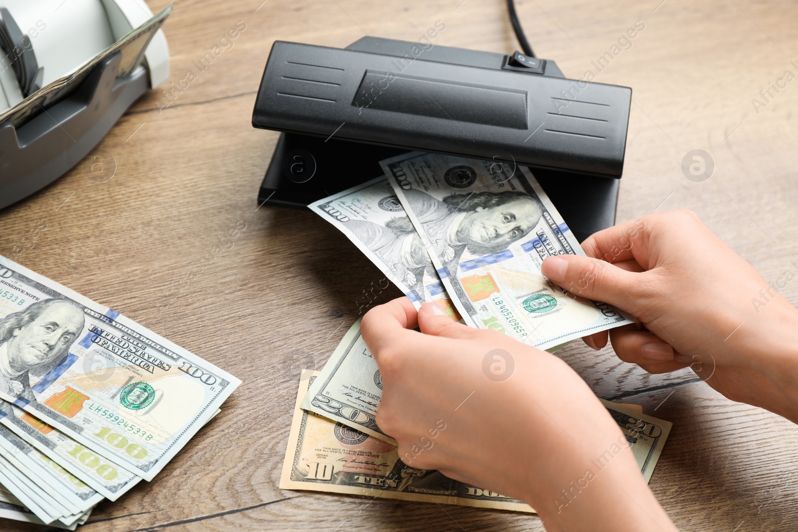 Photo of Woman checking dollar banknotes with currency detector at wooden table, closeup. Money examination device