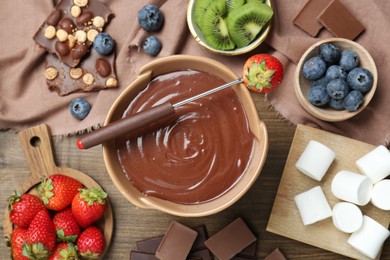 Fondue pot with melted chocolate, marshmallows, fresh kiwi, different berries and fork on wooden table, flat lay