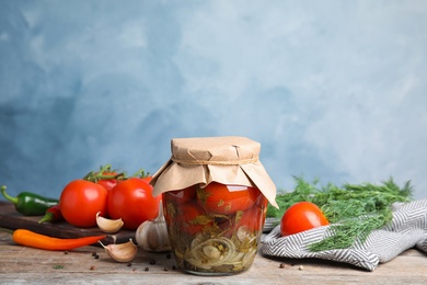 Pickled tomatoes in glass jar and products on wooden table against blue background, space for text