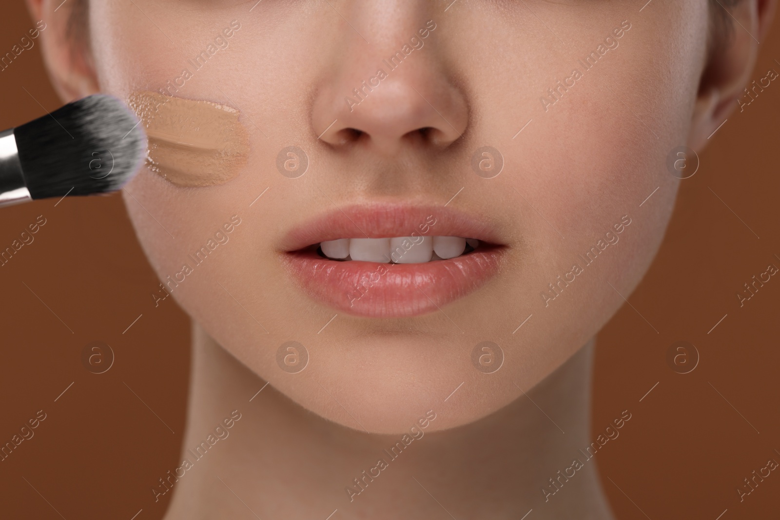 Photo of Teenage girl applying foundation on face with brush against brown background, closeup