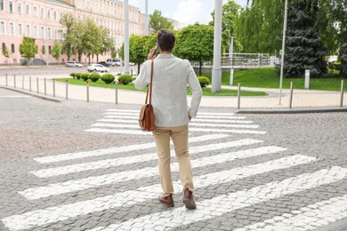 Photo of Young man talking on phone while crossing street, back view. Traffic rules and regulations