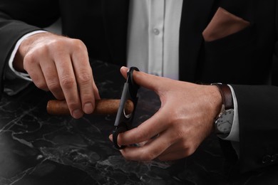 Photo of Man cutting tip of cigar at dark marble table, closeup