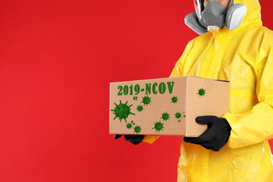 Image of Man in chemical protective suit holding cardboard box on red background, closeup view with space for text. Coronavirus outbreak