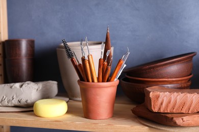 Photo of Set of different crafting tools and clay dishes on wooden rack in workshop, closeup